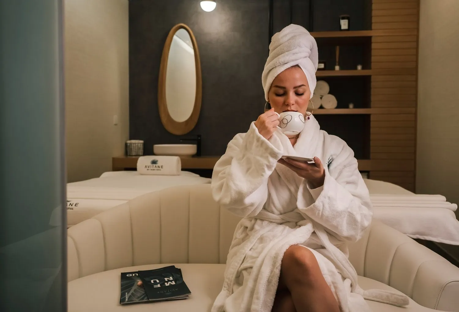 A woman in a robe and hair towel relaxing with a cup of tea in a spa lounge